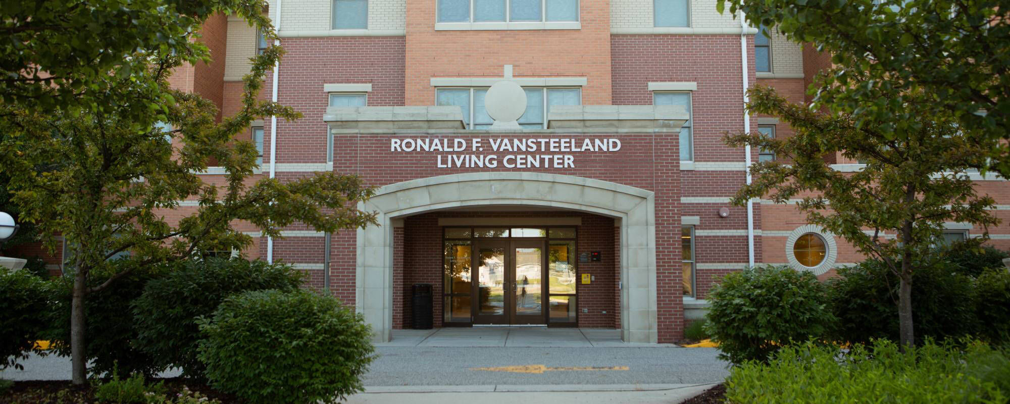 Front view of Ronald F. Vansteeland Living Center, a brick building with an arched entrance, surrounded by green trees and shrubs, conveying a welcoming atmosphere.